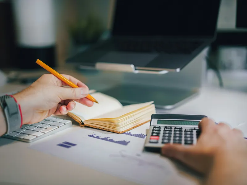 Mains tenant un crayon au-dessus d'un carnet, à côté d'une calculatrice